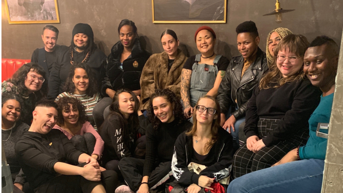 17 mostly BIPOC young people of different genders, skin tones, and sizes are pictured indoors. They are seated close to each other, with some on the floor and others on banquette seating. Some are mid-smile, others are laughing, and everyone is having a good time.